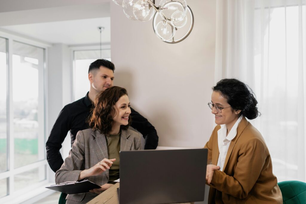 A financial advisor sitting at a table with two clients, reviewing documents and discussing a business plan together in a professional office setting.